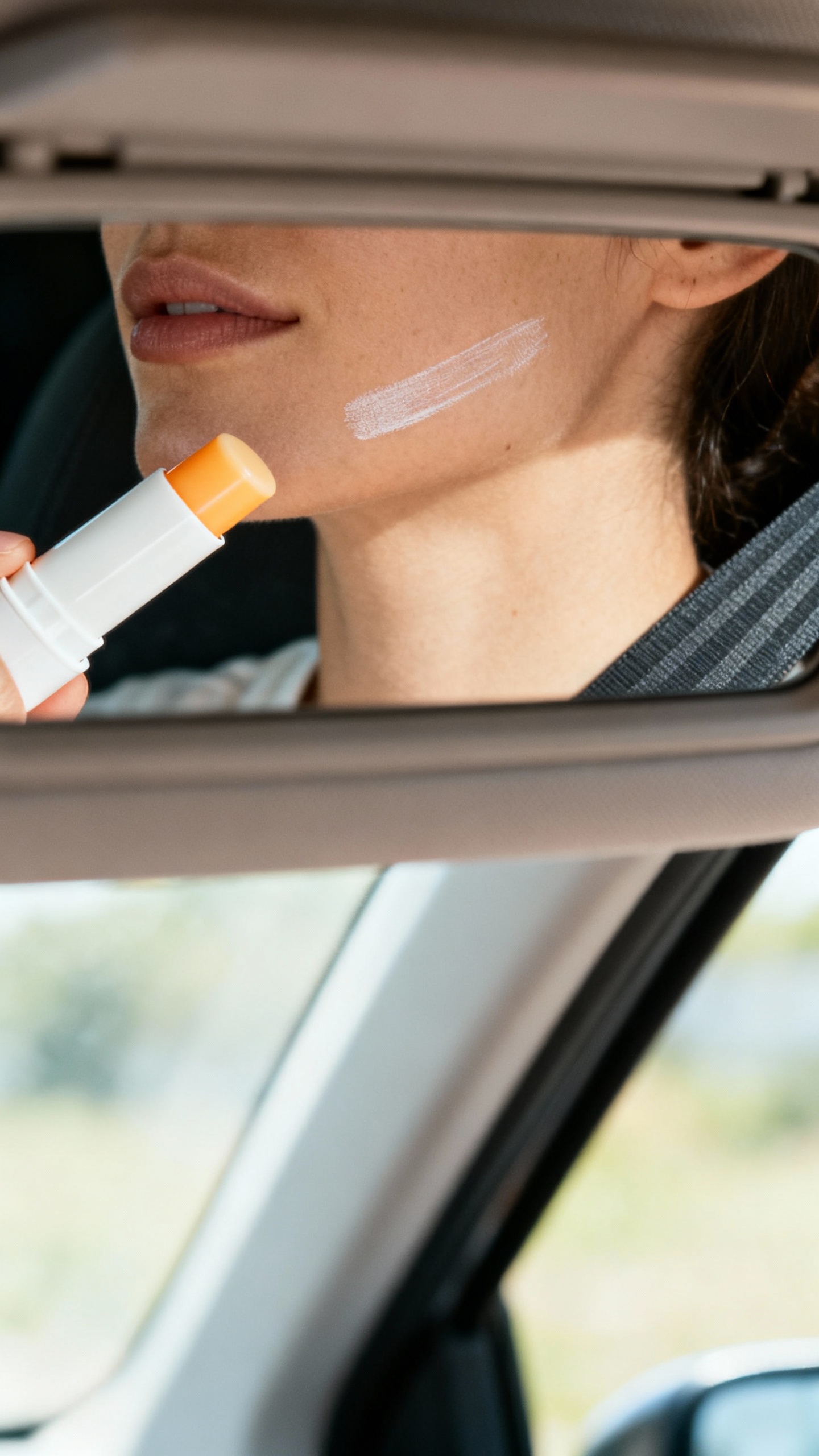 Car visor mirror closeup, sunscreen stick swiped on cheek, daylight, seatbelt visible