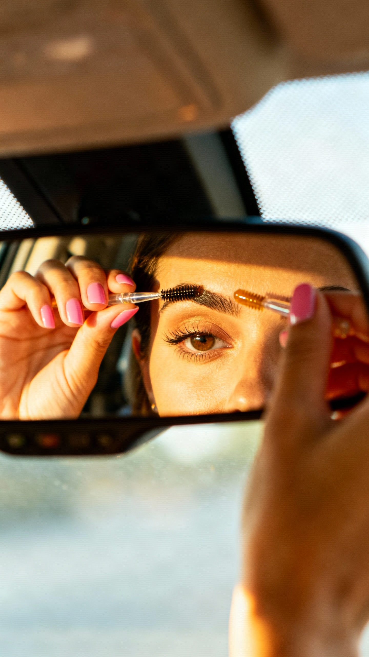 Female hands brushing brows with tinted gel, car rearview mirror reflection