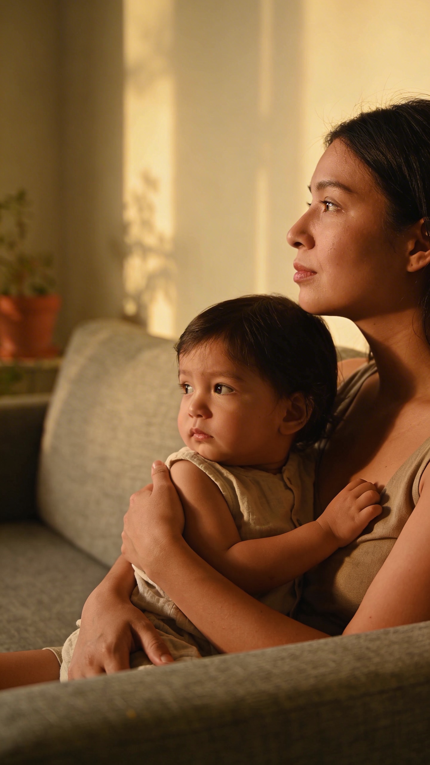 Mom holding toddler, distant gaze on couch, natural light
